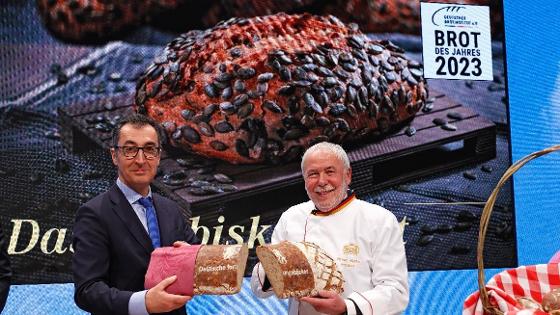 Bundesminister Cem Özdemir und Michael Wippler, Vorsitzender des Zentralverbandes des Bäckerhandwerks, halten ein angeschnittenes Kürbiskernbrot in die Kamera. 