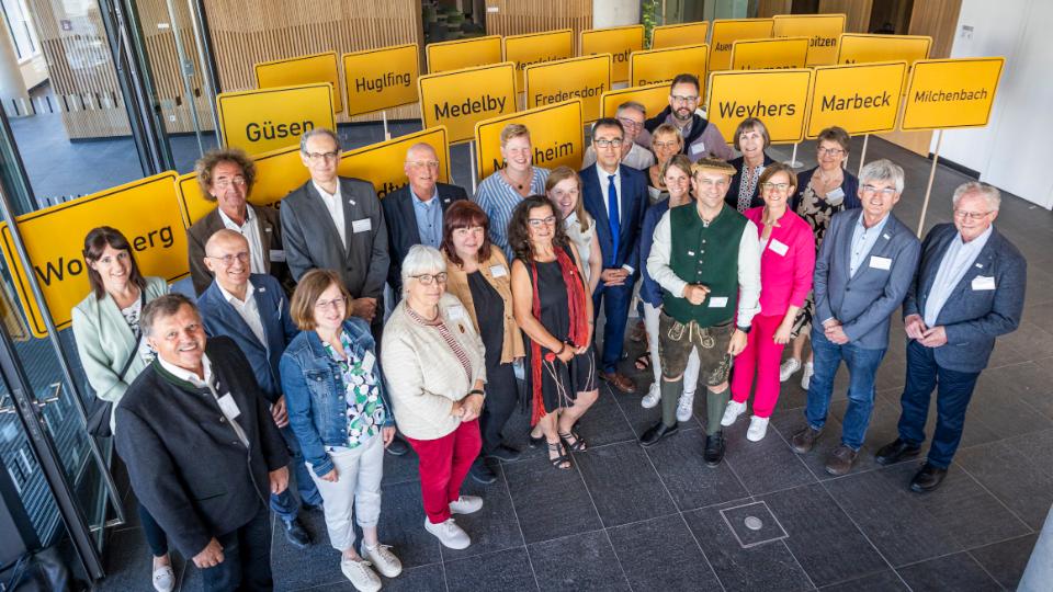 Gruppenfotot Bundesminister  Özdemir mit der Bewertungskommission für den Bundeswettbewerb "Unser Dorf hat Zukunft" im Hintergrund die Ortsschilder der teilnehmenden Gemeinden