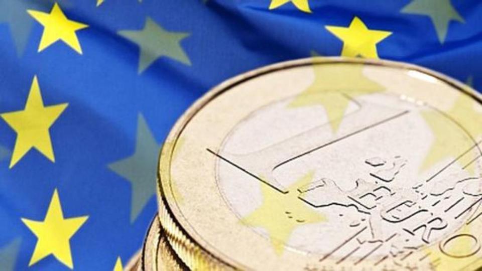 Ein Stapel 1-Euro-Münzen; im Hintergrund die Flagge der Europäischen Union