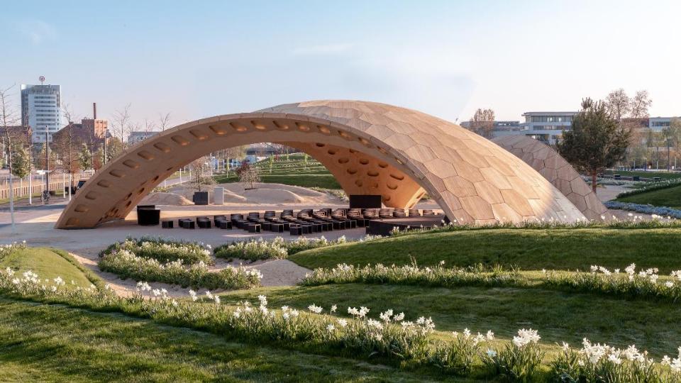 Pavillon aus Holz, darunter eine Bühne und Sitzgelegenheiten. Drumherum kleine Hügel mit Rasen und weißen Blumen.dazwischen eine Grünfläche