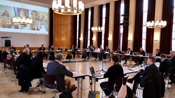 Gruppenbild: Mitglieder der Zukunftskommission Landwirtschaft sitzen in einem großen Raum an Tischen, im Hintergrund weitere Personen auf einer Leinwand per Video zugeschaltet