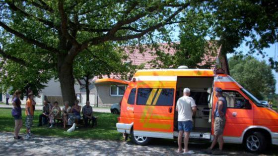 Ein roter VW-Bus steht unter einem Baum; daneben stehen und sitzen wartende Menschen.