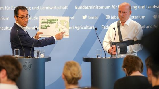 Bundesminister Cem Özdemir und Prof. Pablo Steinberg, Präsident des Max Rubner-Instituts stehen an Rednerpulten, der Bundesminister hält eine Tafel mit Informationen über den Zukergehalt von Kindergetränken in die Kamera, 