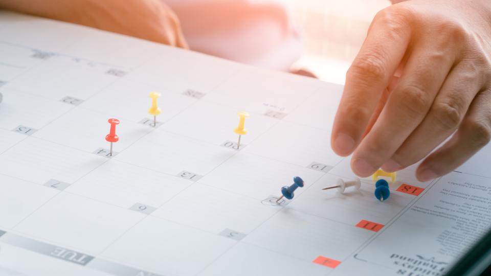 Hand steckt Pins auf einen Kalender; daneben steht ein Laptop