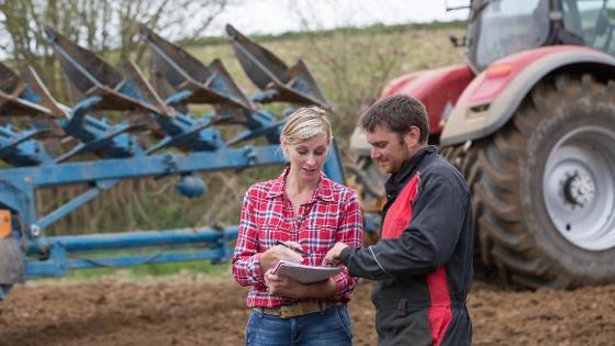 ein Mann und eine Frau stehen auf dem Acker im Gespräch über ein Papier gebeugt, im Hintergrund ein Traktor