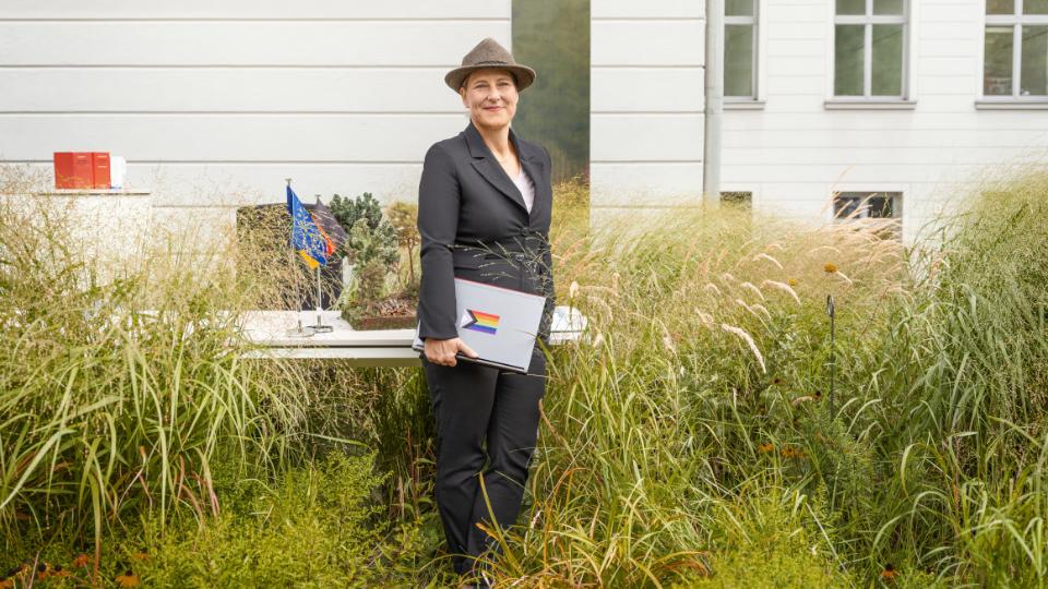 Eine Mitarbeiterin des BMEL mit grünen Försterhut hält einen Laptop mit LGBTQ+-Aufkleber in der Hand und steht vor einem Schreibtisch, um sie herum grüne Pflanzen. Im Hintergrund protokollarische Flaggen von Deutschland und der EU sowie Bücher. 