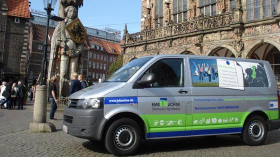 Ein Kleinbus mit Ems-Achse-Werbung steht auf dem Bremer Marktplatz; im Hintergrund der Bremer Roland. 