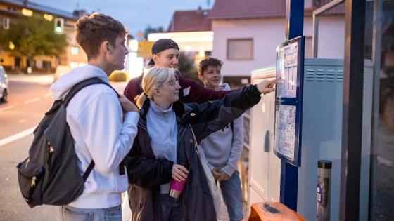 Jugendliche stehen an der Bushaltestelle und betrachten den Fahrplan