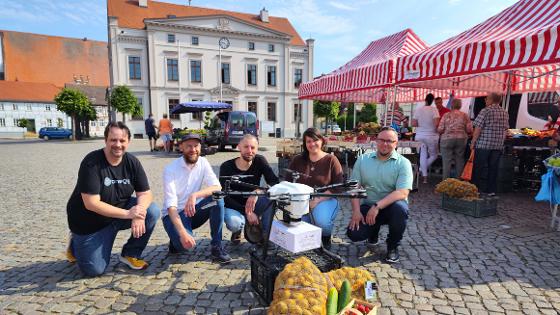 Das Projektteam des Projekts "Stadt-Land-Drohne" kniet auf dem Marktplatz in Wusterhausen hinter eine Drohne, davor zwei Säcke Kartoffeln, Gurken und ein Korb mit Erdbeeren, im Hintergrund das Rathaus