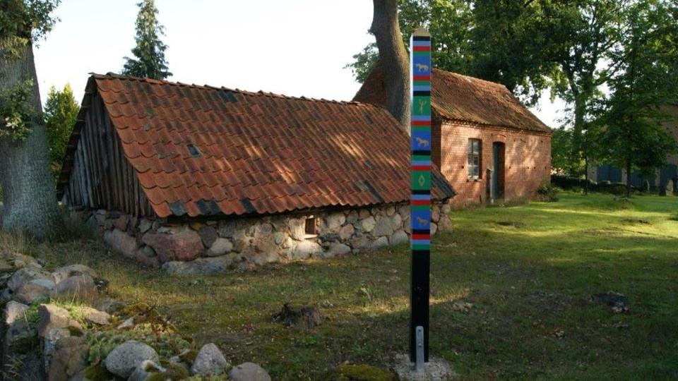 Haus aus Feldsteinen und Backsteinhaus auf einer Wiese mit einem bemalten Pfosten im Vordergrund Haus aus Feldsteinen und Backsteinhaus auf einer Wiese mit einem bemalten Pfosten im Vordergrund