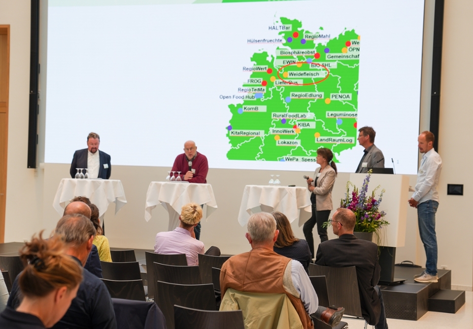 Ein Fachkongress zum Thema "Erfolgreiche Regionalvermarktung". 5 Personen stehen auf der Bühne und stellen beispielhaft Projekte der Regionalvermarktung vor. Im Hintergrund ist eine Landkarte zu sehen. Ein Fachkongress zum Thema "Erfolgreiche Regionalvermarktung". 5 Personen stehen auf der Bühne und stellen beispielhaft Projekte der Regionalvermarktung vor. Im Hintergrund ist eine Landkarte zu sehen.
