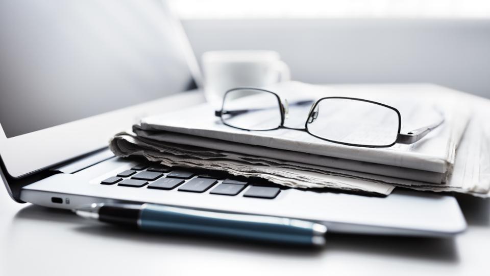 Newspapers and eyeglasses are laying on a laptop, a pen is next to it