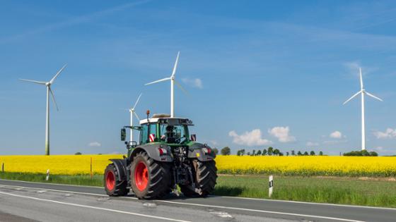 A tractor is driving on a road. In the background is a canola field and wind power plants