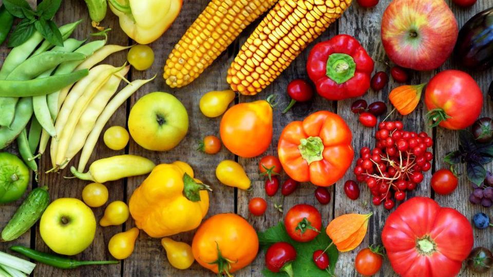 Various types of fruit and vegetables are arranged next to each other sorted by colour: from green beans and cucumbers to yellow apples and peppers to red tomatoes and finally blue berries and eggplant.